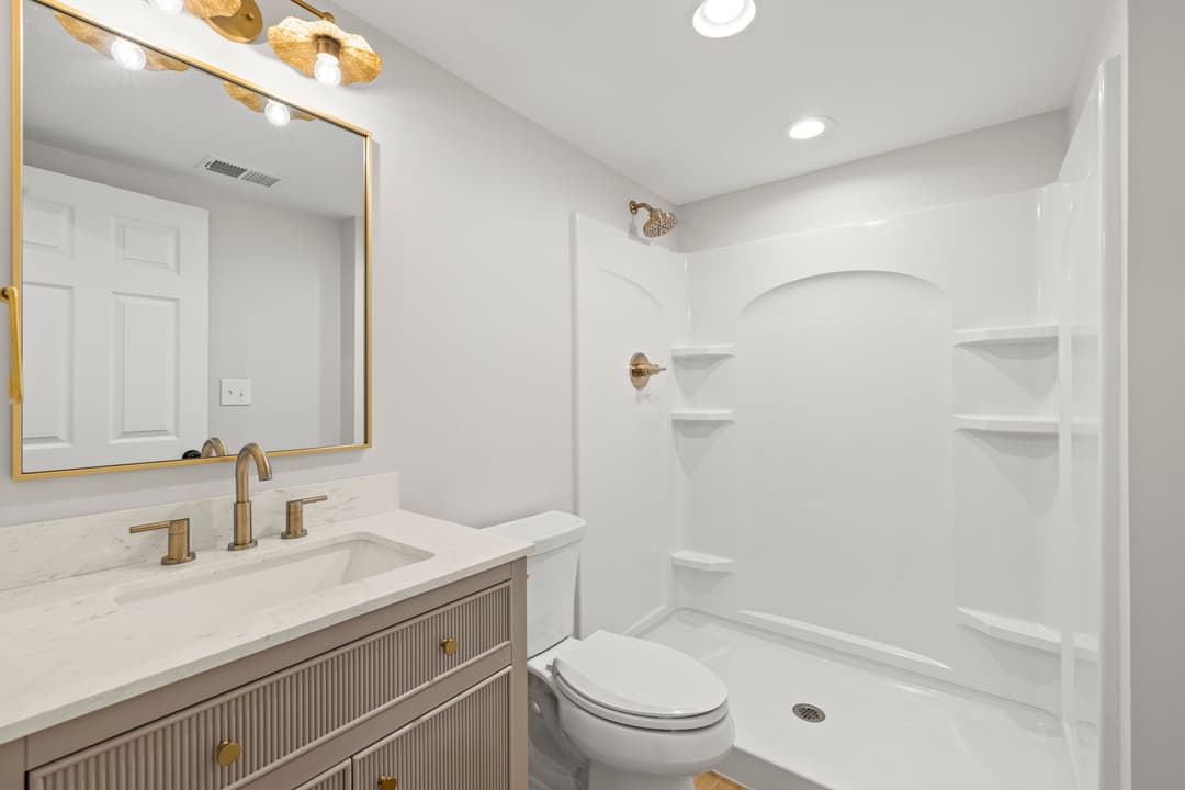 Modern bathroom with light fixtures, gold faucet, and white shower stall. Elegant mirror above sink.