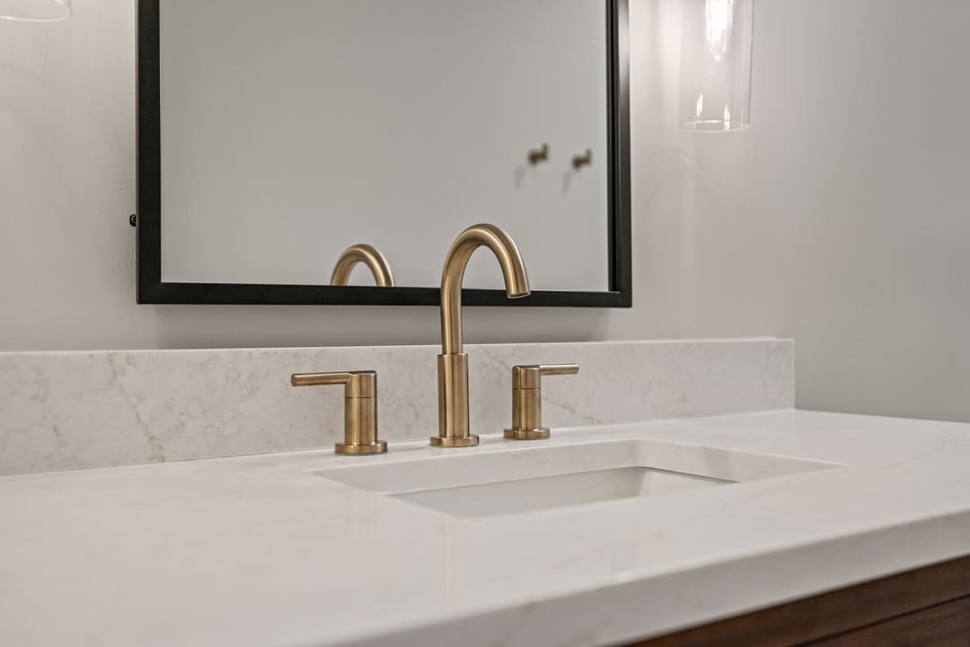 Modern bathroom sink with gold faucet and marble countertop, featuring a large mirror above.