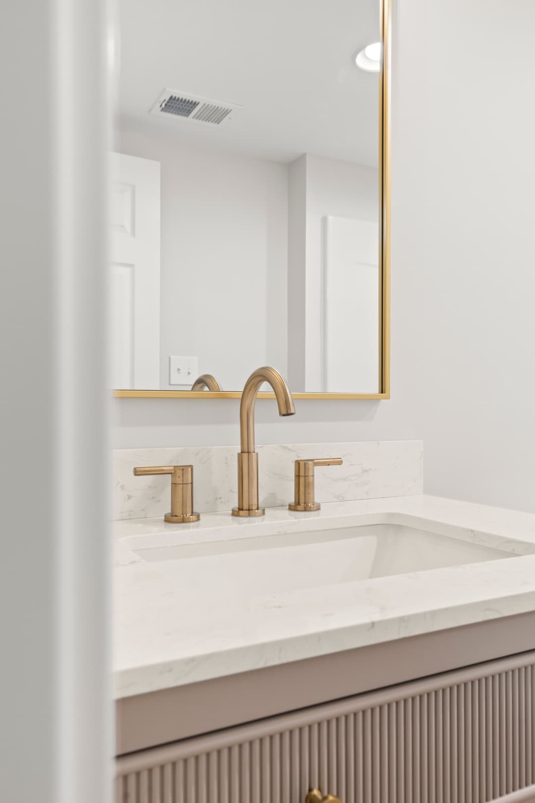 Modern bathroom sink with gold faucets and a large wall mirror against a light backdrop.