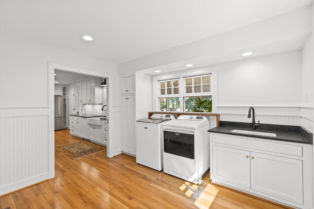 Modern laundry room with white appliances, black sink, and hardwood floors in bright space.