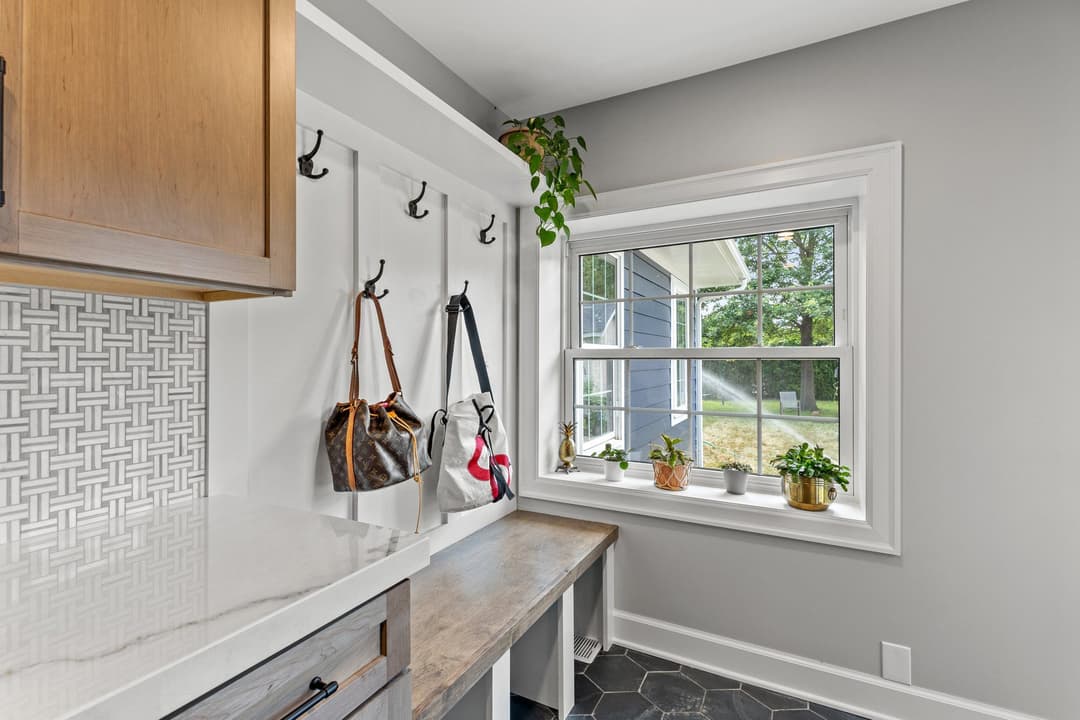 Modern mudroom with hooks for bags, a wooden bench, and potted plants by a sunny window.