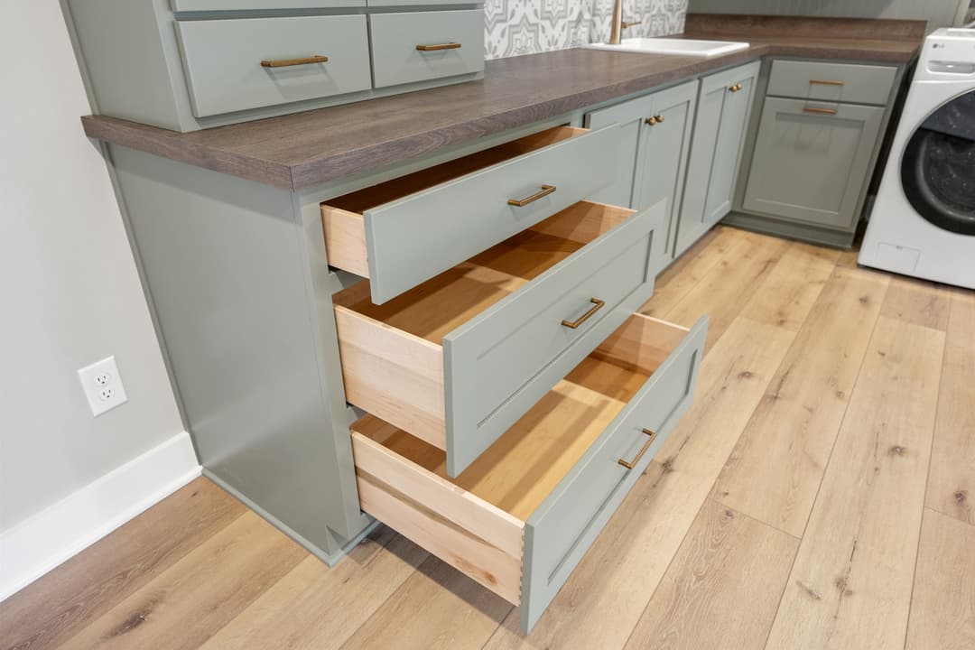 Modern light green kitchen cabinet with open drawers showcasing wooden interiors and a laundry area.