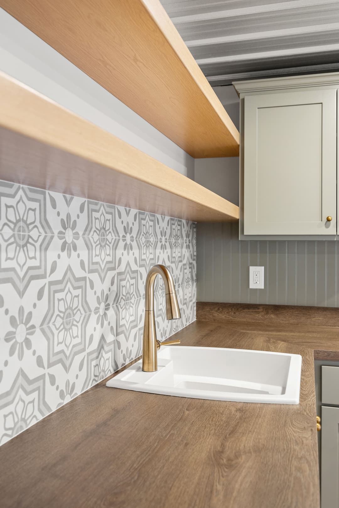 Modern kitchen with gray patterned backsplash, wooden shelves, and stylish gold faucet.